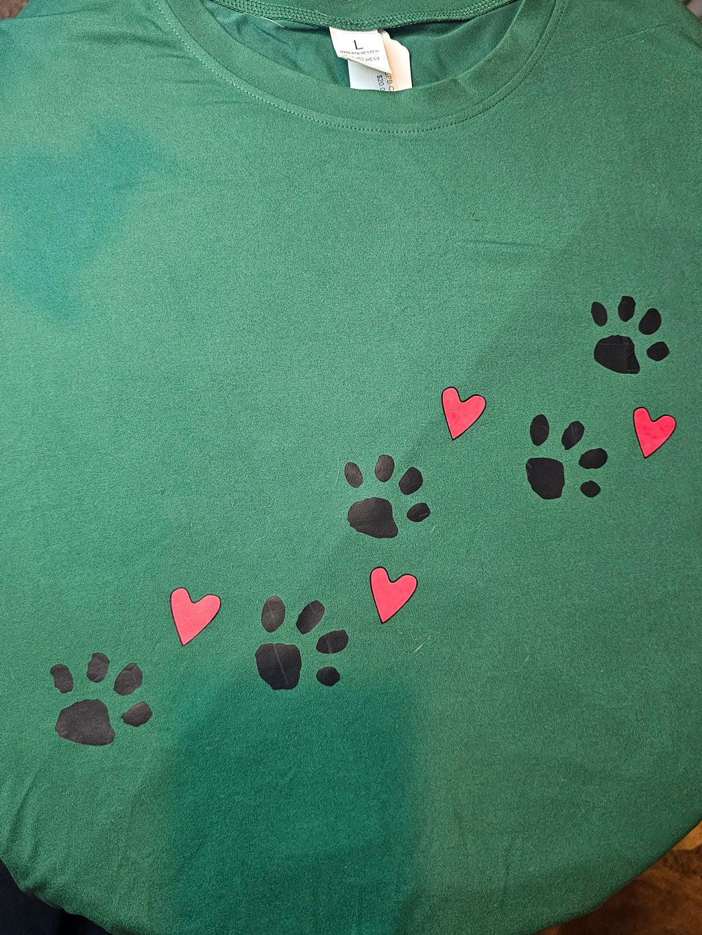 A green crewneck t-shirt laid flat, featuring a printed design of black paw prints and pink hearts arranged diagonally across the chest.