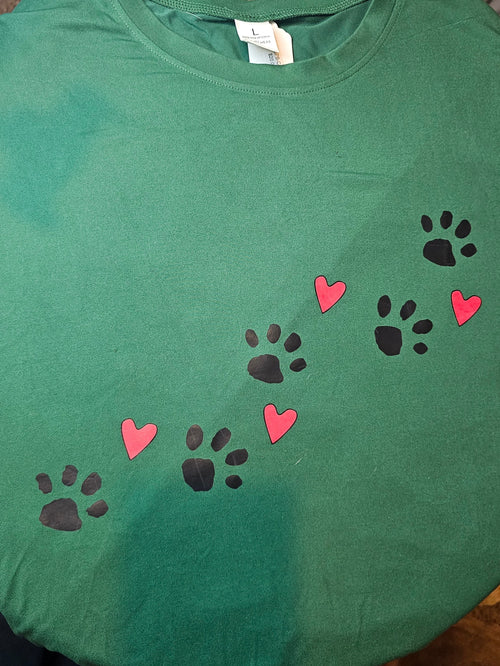 A green crewneck t-shirt laid flat, featuring a printed design of black paw prints and pink hearts arranged diagonally across the chest.