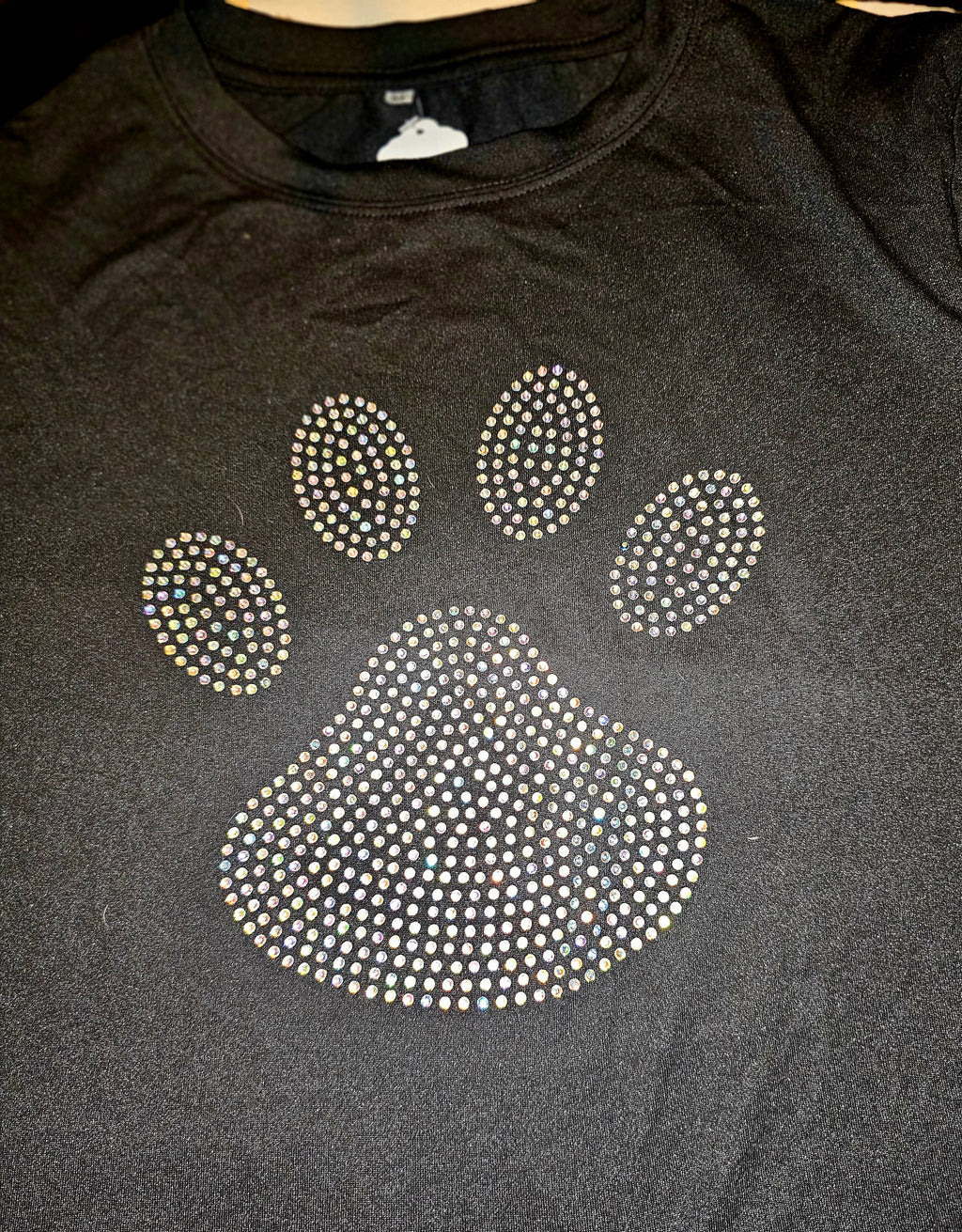 A black crewneck sweatshirt featuring a large paw print graphic embellished with sparkling rhinestones, laid flat on a dark surface.