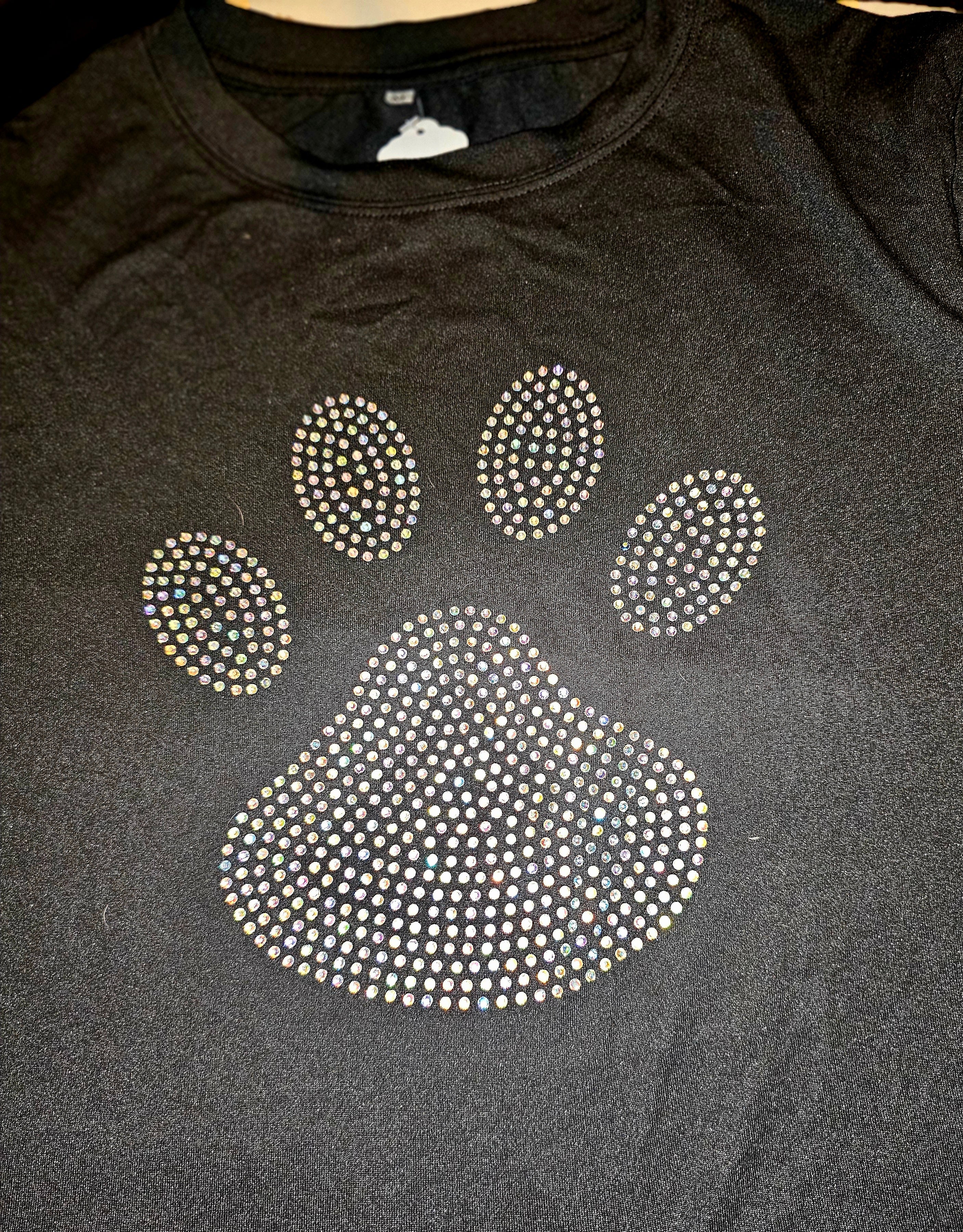 A black crewneck sweatshirt featuring a large paw print graphic embellished with sparkling rhinestones, laid flat on a dark surface.